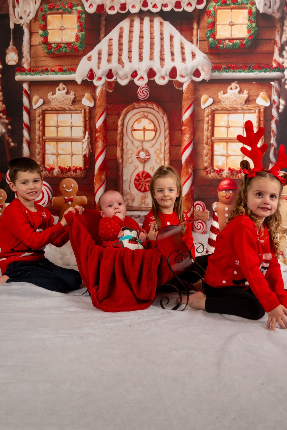 Children in red outfits sitting in front of gingerbread house backdrop for Christmas photo.