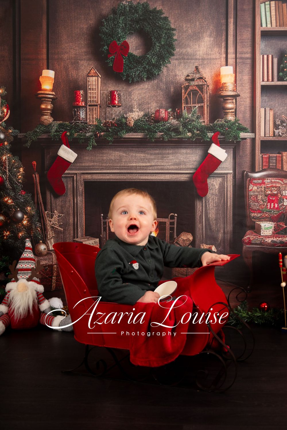 Excited baby in black outfit sitting in red chair by Christmas decorated fireplace.