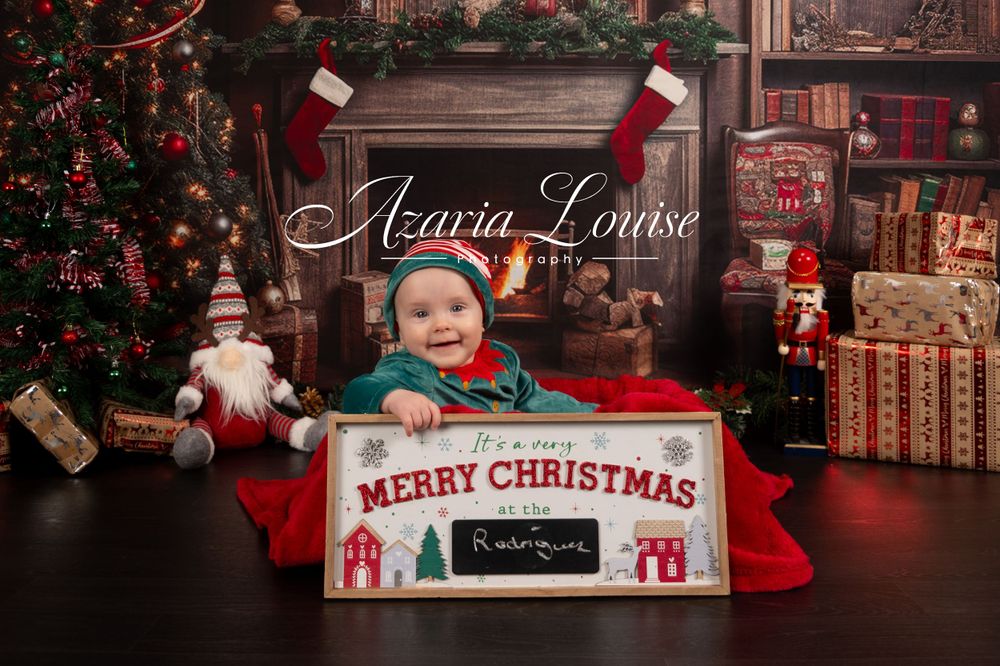Baby in festive attire poses with Merry Christmas sign in cozy holiday setting.