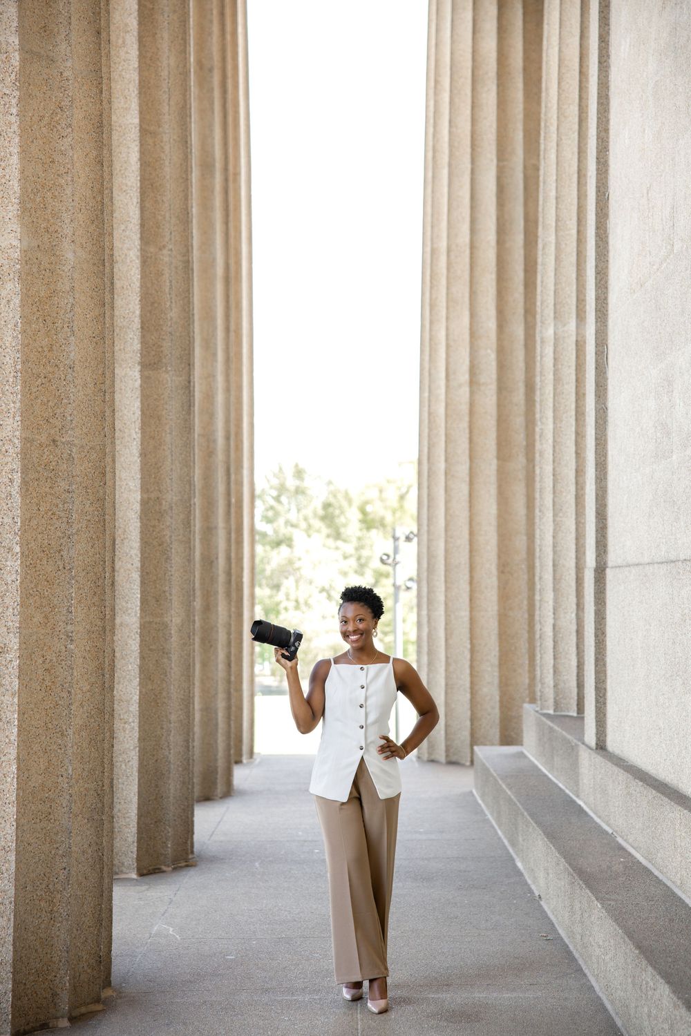 Professional wearing a beige pantsuit and white top stands confidently between classic stone columns with a handheld device.