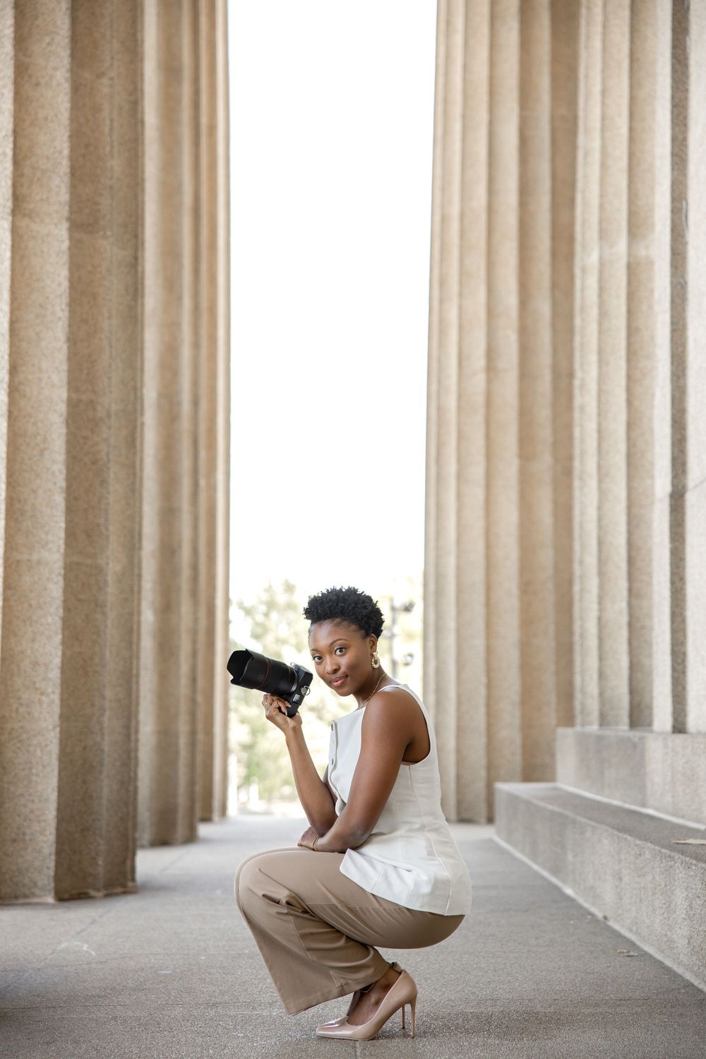 Professional photographer crouching with camera between classic stone columns in elegant business attire.