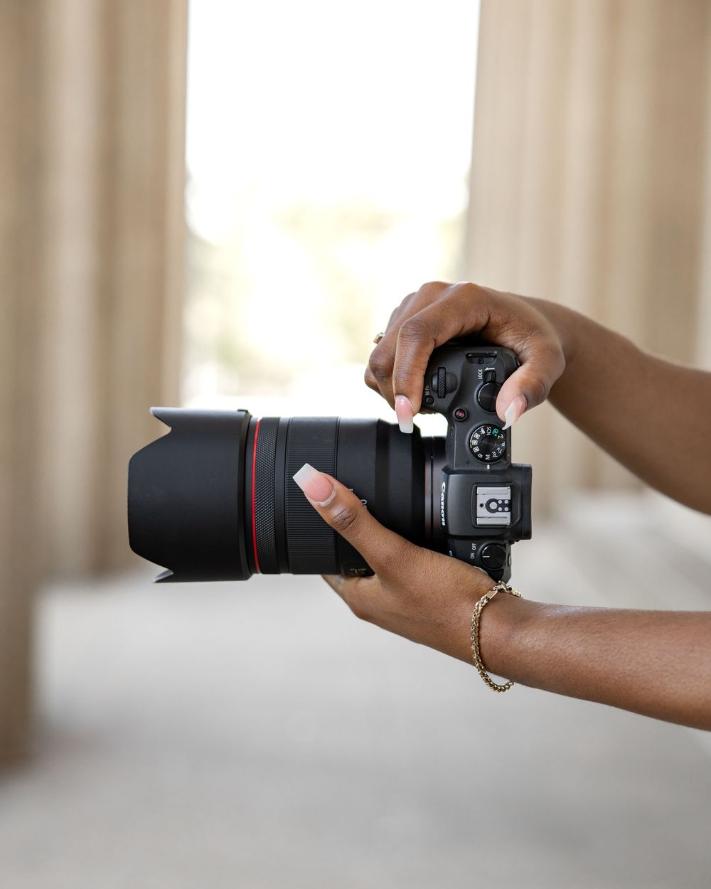 Hands holding a professional DSLR camera with telephoto lens against a bright window background.