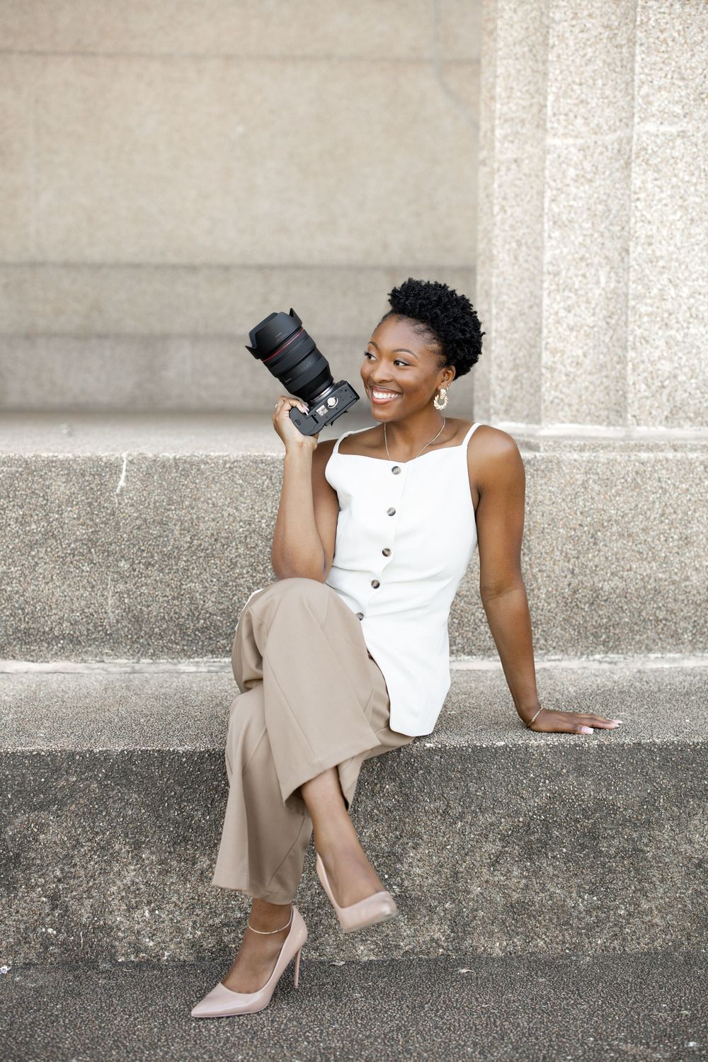 Woman in white sleeveless top and beige skirt sits on concrete steps while holding a drink and smiling.