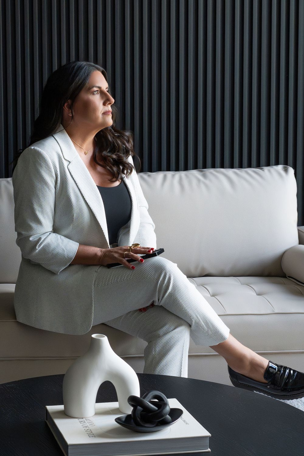 Person in white suit sitting on couch with phone against dark striped wall background.