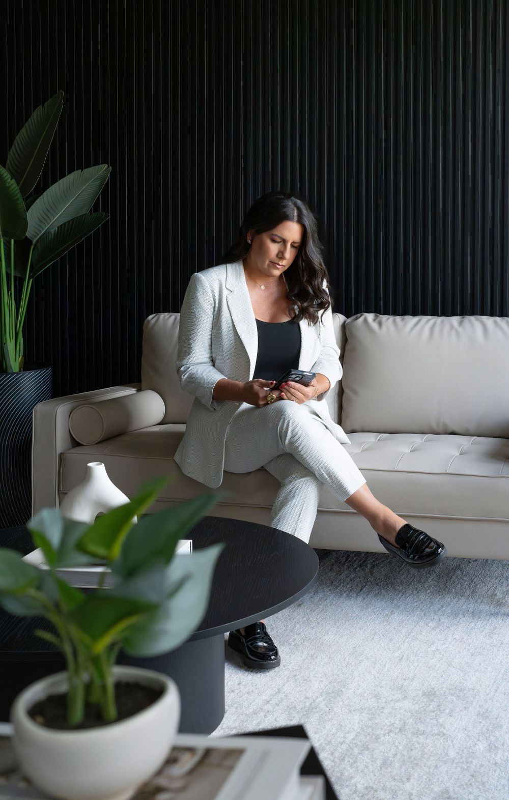 Person in white suit relaxing on white couch with phone near modern indoor plants.