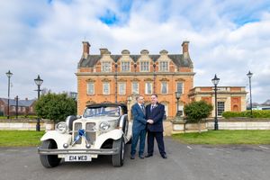 A classic white vintage wedding car parked in front of a brick mansion with cloudy blue skies overhead. Casual Captures Wedding Photography
