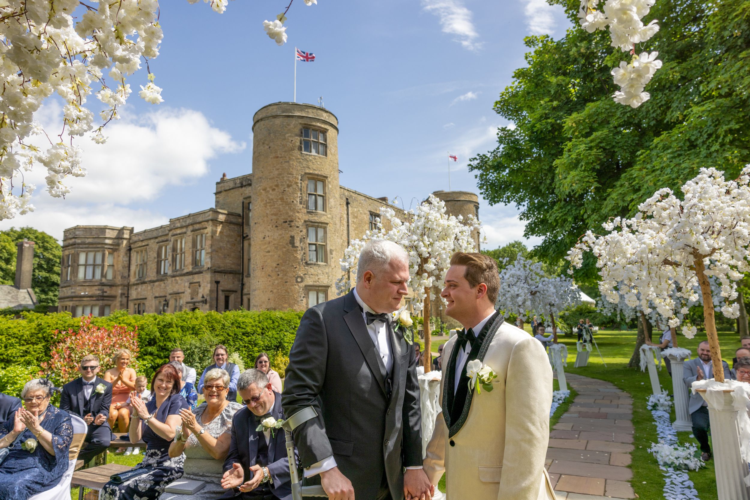 A wedding ceremony takes place outdoors at a historic castle with cherry blossoms. Casual Captures Wedding Photography