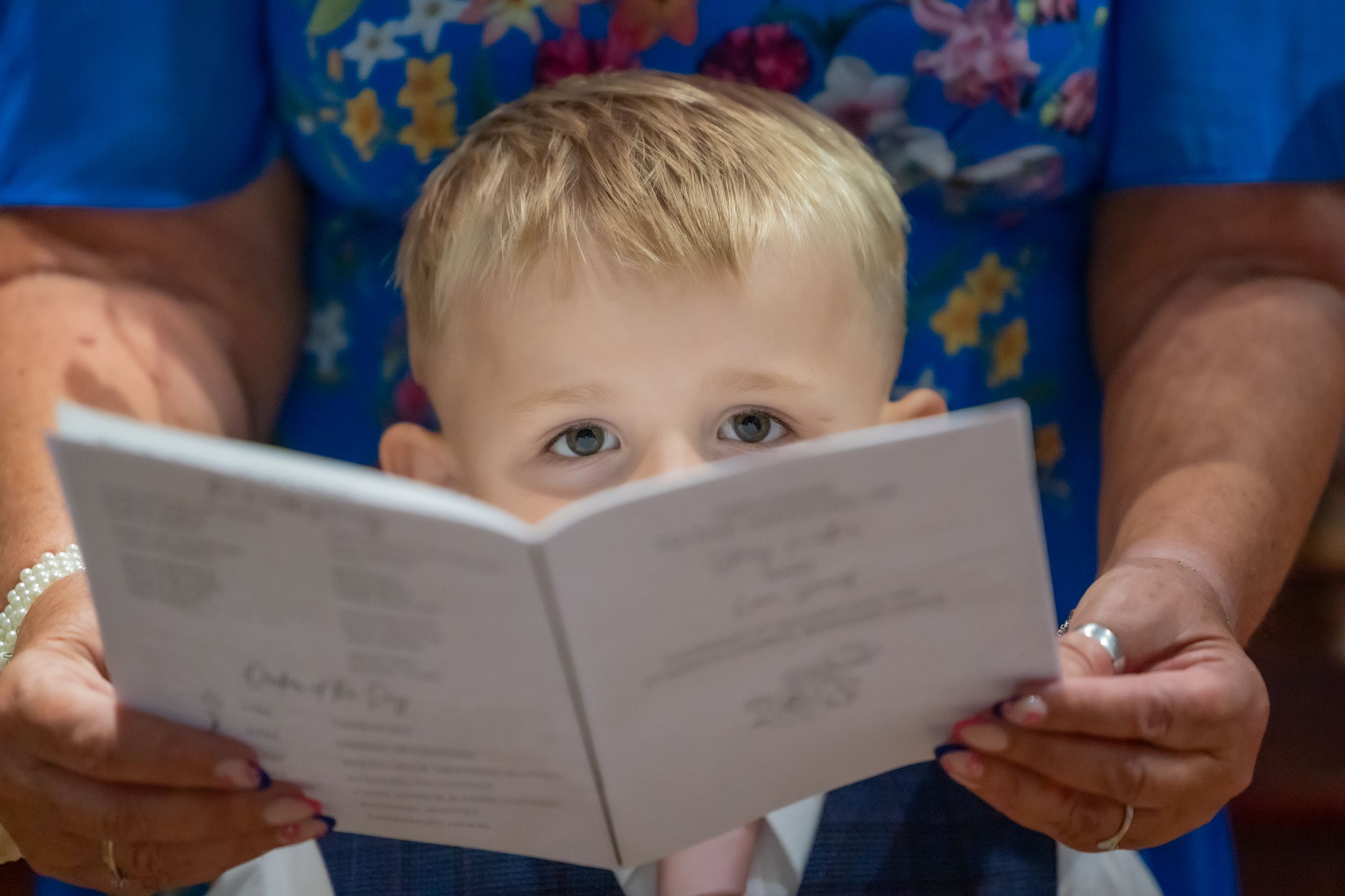 Small child peering over an open book being held by adult hands wearing a blue shirt.