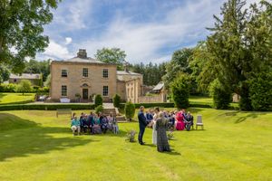 Outdoor wedding ceremony on lush green lawn before elegant Georgian country house on a sunny summer day.