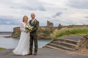 A wedding couple poses along the scenic coastal cliffs of Tyneside with castle ruins and ocean views in the background.