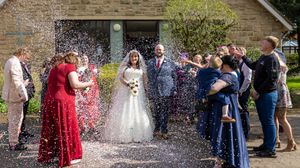 Wedding guests celebrate by throwing confetti over newlyweds outside a stone building on their special day.