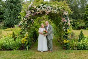 Couple stands under floral arch in lush garden wedding ceremony, bride holds bouquet, groom in gray suit.