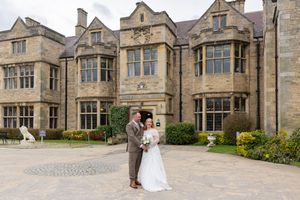 A couple stands on a stone driveway in front of a grand English manor house with Gothic architecture and bay windows.