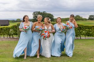 Wedding party in matching light blue dresses poses outdoors in a vineyard setting with coral bouquets.