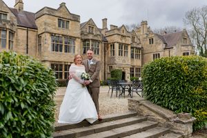 A couple stands on stone steps in front of an elegant historic manor house with Gothic architectural details.