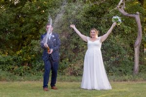 Bride cheers as groom pops champagne bottle outdoors at wedding celebration surrounded by lush green trees.