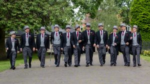 A line of groomsmen in matching dark suits and red ties walk along a garden path.