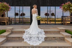 Bride in elegant backless wedding gown with lace train stands on stone steps at a countryside venue.
