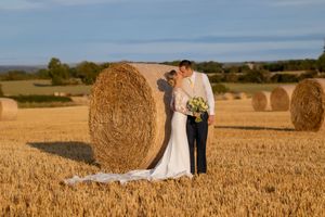 Bride and groom embracing beside a large hay bale in a golden wheat field on their wedding day.