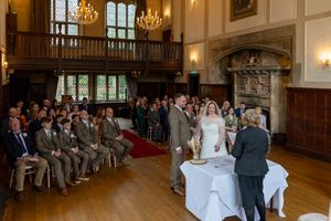 Wedding ceremony taking place in an elegant historic hall with wood paneling, chandeliers and guests seated in rows.