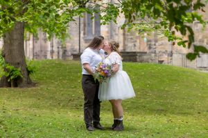 Two people embrace in a romantic moment on a lawn surrounded by trees and historic stone buildings. Casual Captures Wedding Photography