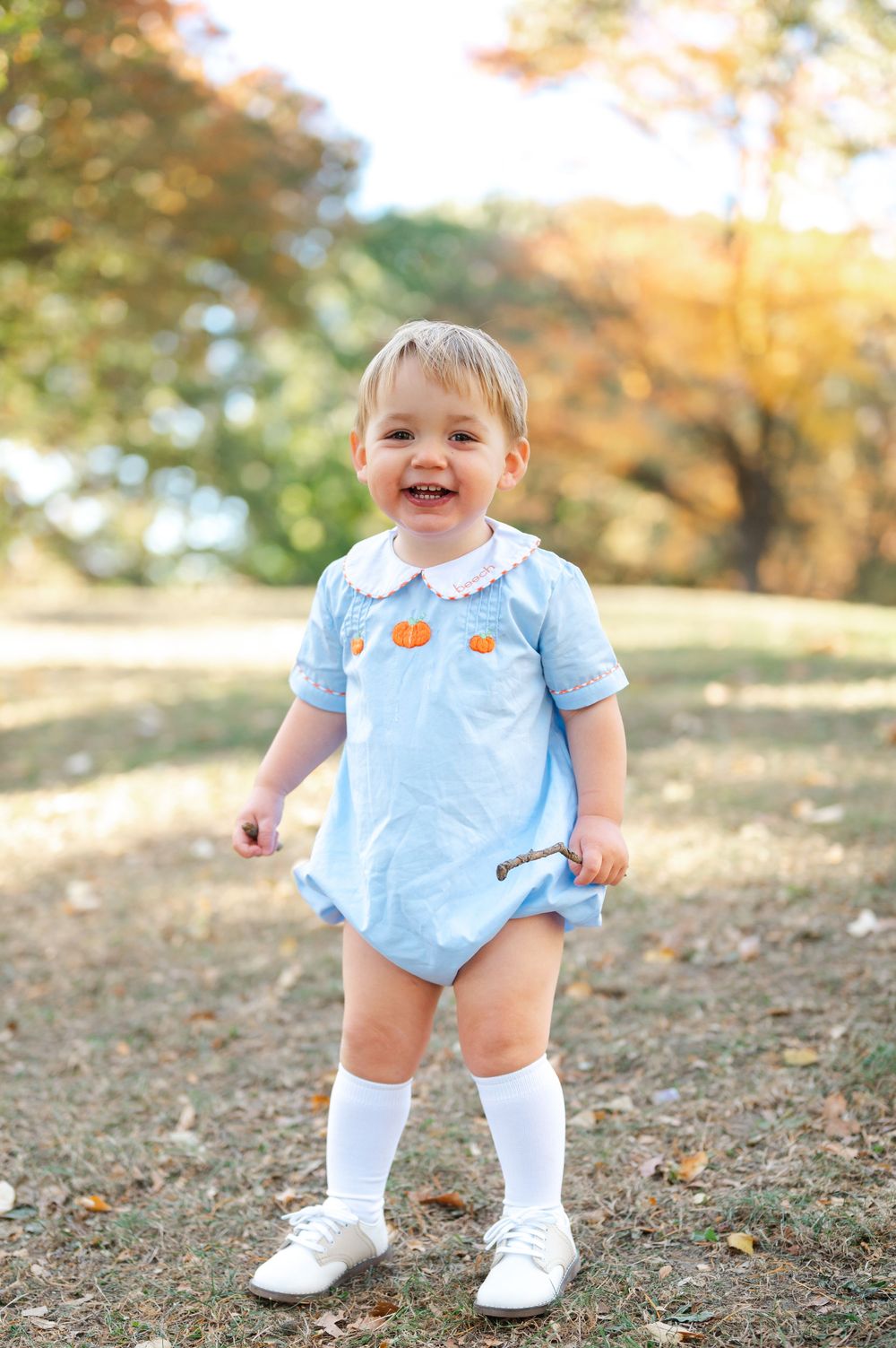 A smiling toddler wearing a light blue romper with pumpkin details and white knee socks stands in a sunny autumn setting.