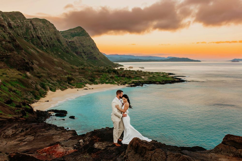 Couple in white embraces on dramatic coastal cliffs at sunset overlooking turquoise Hawaiian waters.