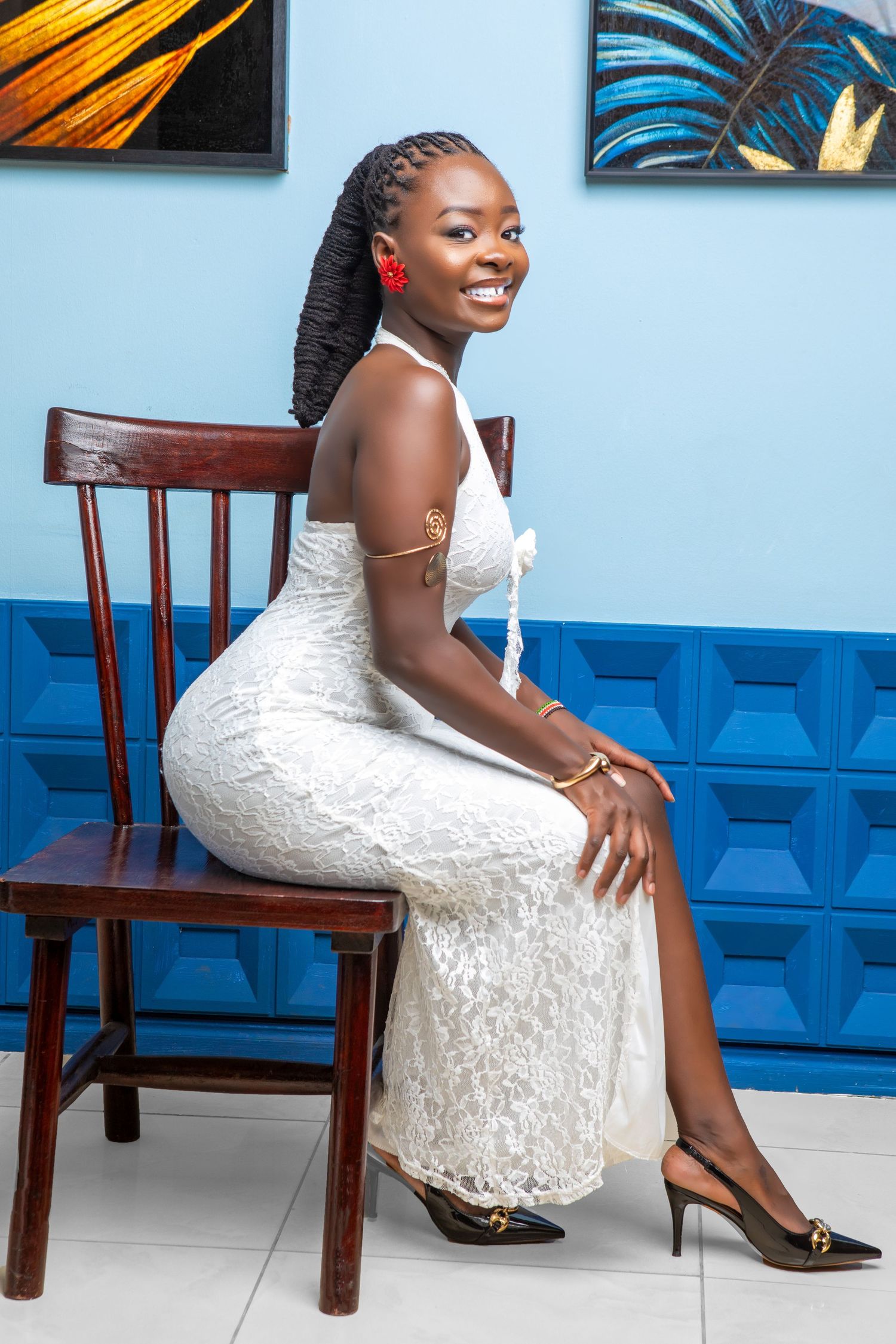 A person in a white lace dress poses elegantly on wooden chairs against a blue and white wall background.