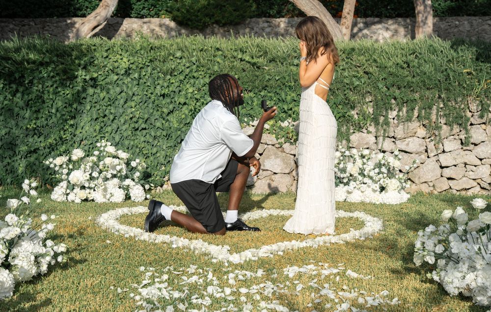 A person in white clothing walks along a stone wall with cascading white flowers and greenery in a garden setting.