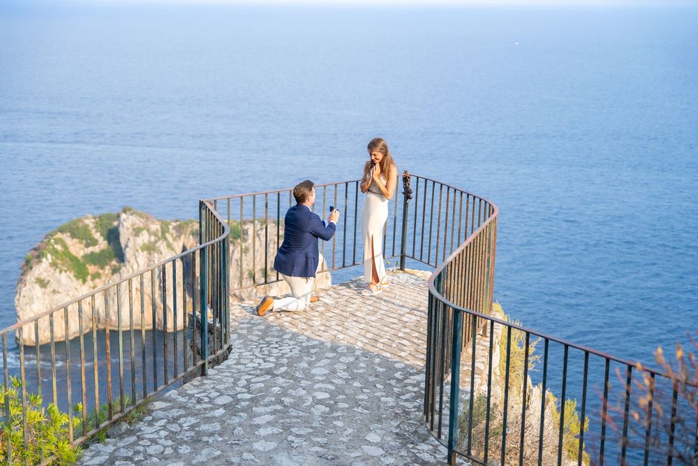 A romantic proposal moment unfolds on a scenic stone overlook with curved railings above the blue Mediterranean Sea.