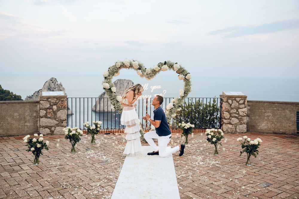 A romantic proposal moment at a scenic coastal overlook with a heart-shaped floral arch and white aisle runner.