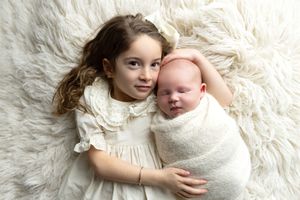 A newborn baby wrapped in soft white fabric lies on a fluffy white rug while being gently embraced.