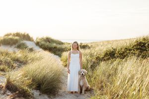 A person in a white dress and their golden retriever dog stand together in tall grass during golden hour.