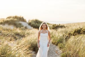 A person in a flowing white dress stands in a golden field at sunset with tall grass swaying in the breeze.