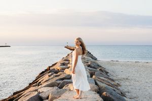 A person in a white dress stands on a rocky jetty extending into the calm ocean at sunset.