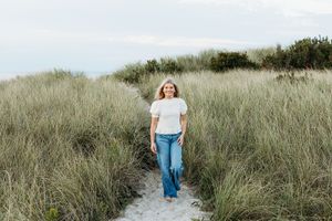 A figure walks along a sandy beach path through tall coastal grass on an overcast day.