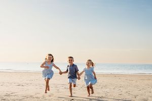 Three people in blue outfits run and hold hands together on a sunny beach with ocean waves in the background.