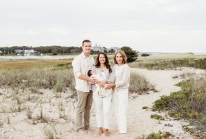 A group of people wearing white outfits pose together on a sandy beach with coastal vegetation in the background.