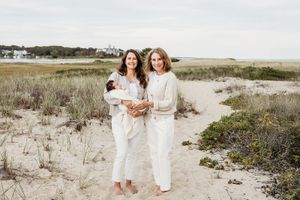 Two people in white outfits walk together on a sandy beach with coastal vegetation and dunes in the background.