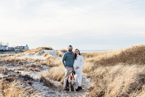 A couple stands with their dog on a coastal path through golden beach grass on an overcast day.