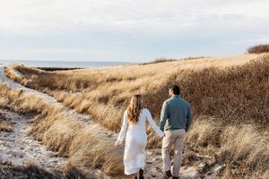 A couple walks along a winding dirt path through golden brown grass fields on an overcast day.