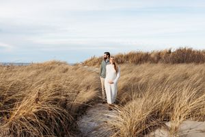 A couple walks through tall golden beach grass along a sandy coastal path on an overcast day.