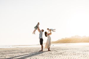 A couple lifts two children playfully on a sunlit beach at sunset, creating joyful silhouettes against the bright sky.