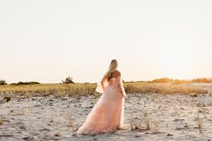 A person in a flowing pink dress walks along a scenic beach shoreline at sunset.