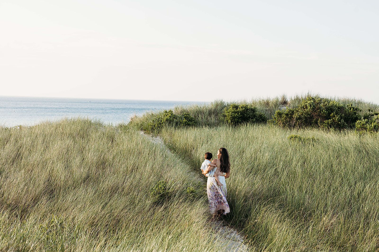 Couple walking together through tall grass on a scenic coastal path with ocean views in the background.