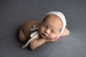 Sleeping newborn baby wearing white knit hat rests peacefully on gray blanket during professional photo session.