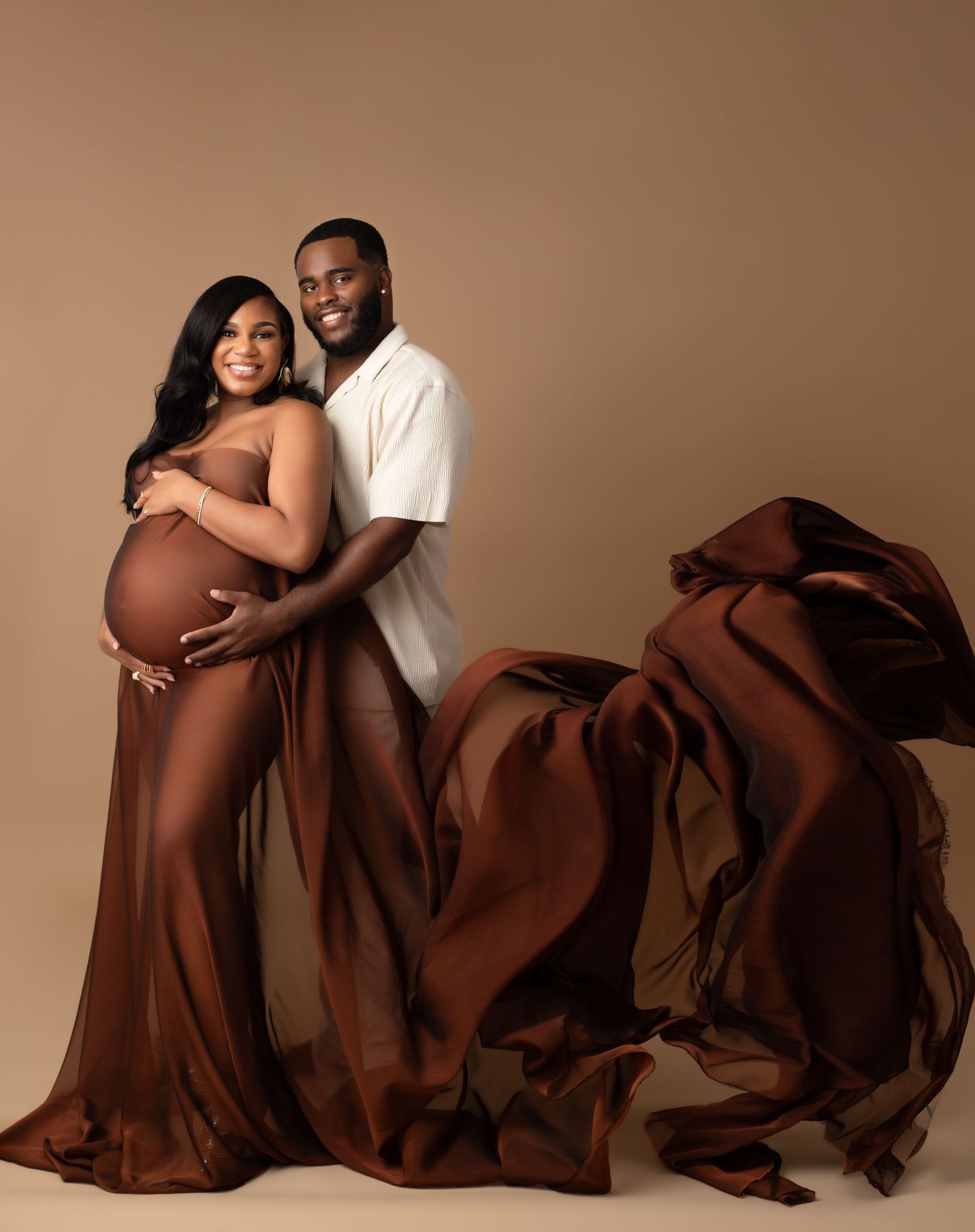 A couple poses together on a brown backdrop while wrapped in flowing chocolate brown fabric creating an artistic maternity photo.