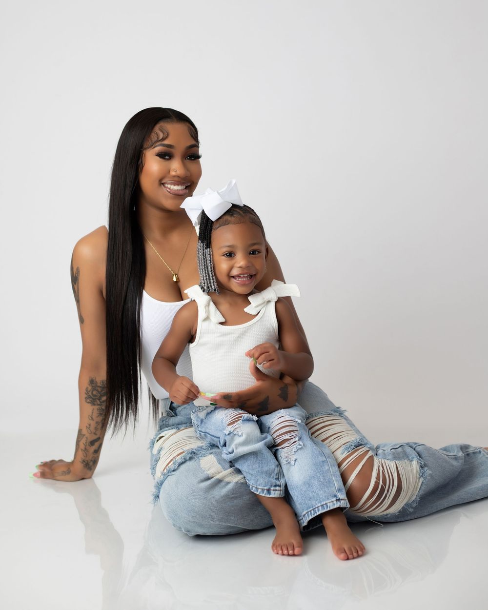 A parent and child wearing matching white tops and distressed jeans pose together on a white backdrop.