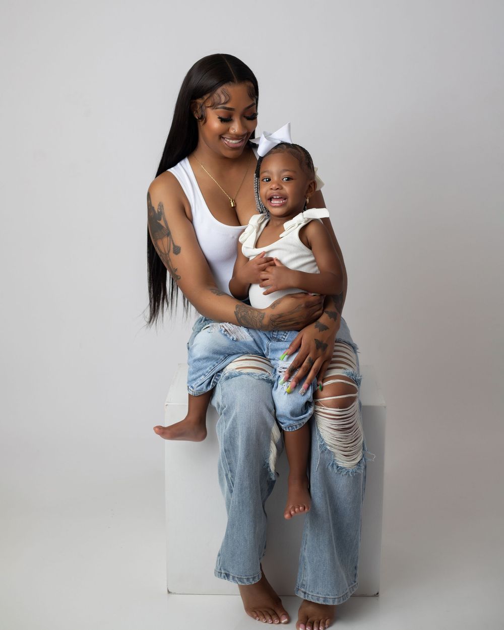 Parent and child sitting together on a white stool wearing casual attire against a gray studio backdrop.