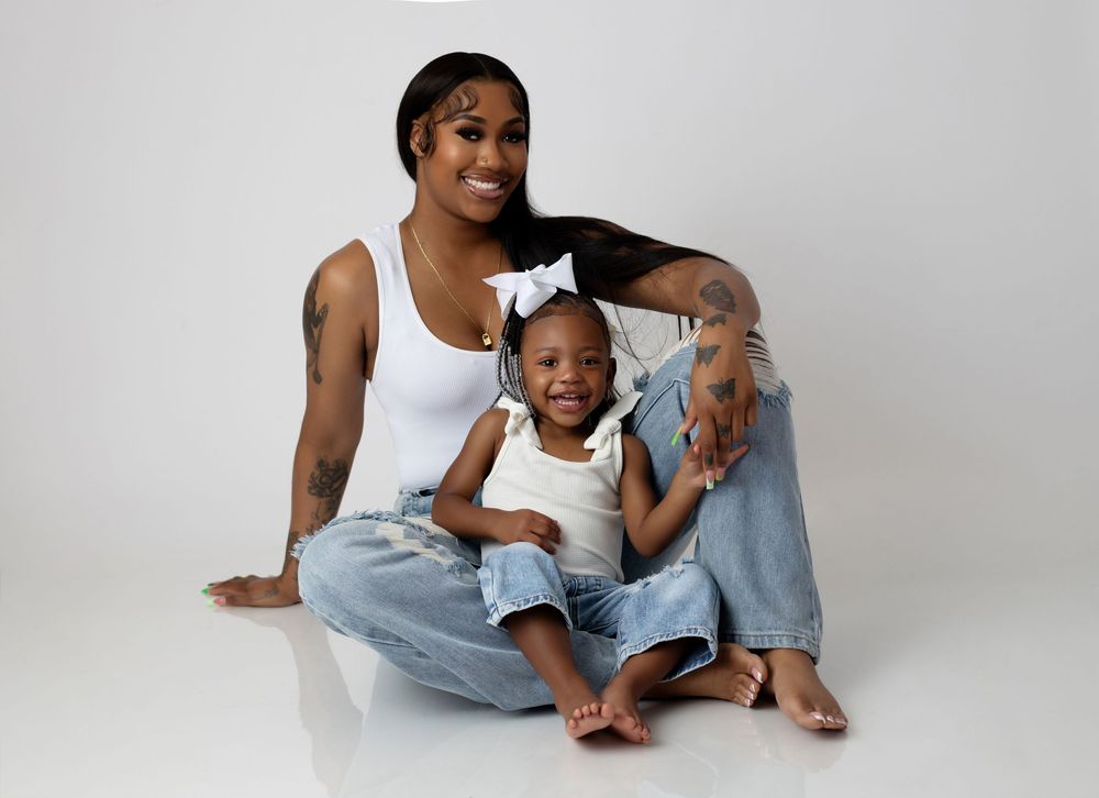 A parent and child wearing white tops and denim sit together on a white backdrop for a casual studio portrait.